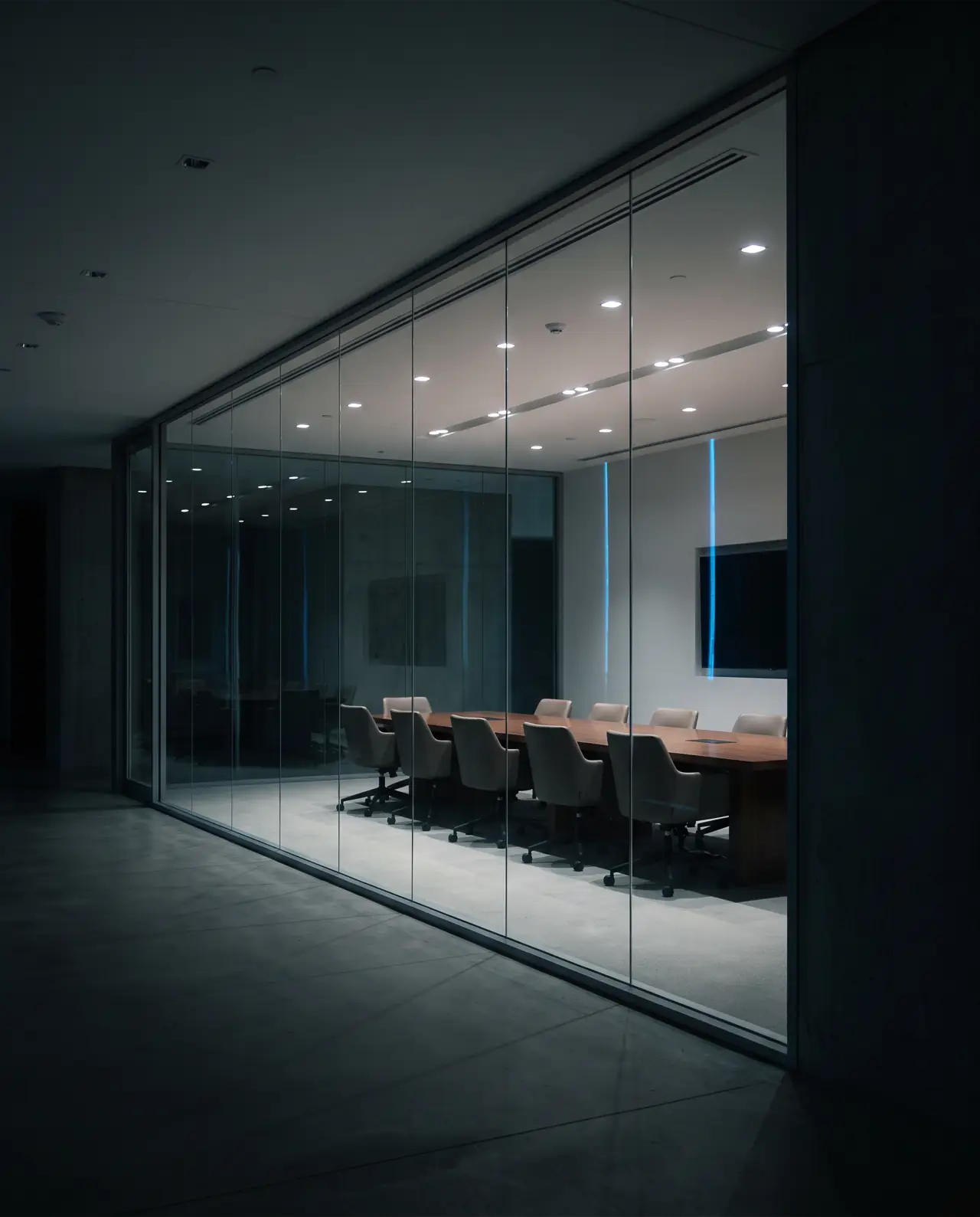 Sala de reunião envidraçada e vazia, acesa por dentro, vista de um corredor escuro à noite
