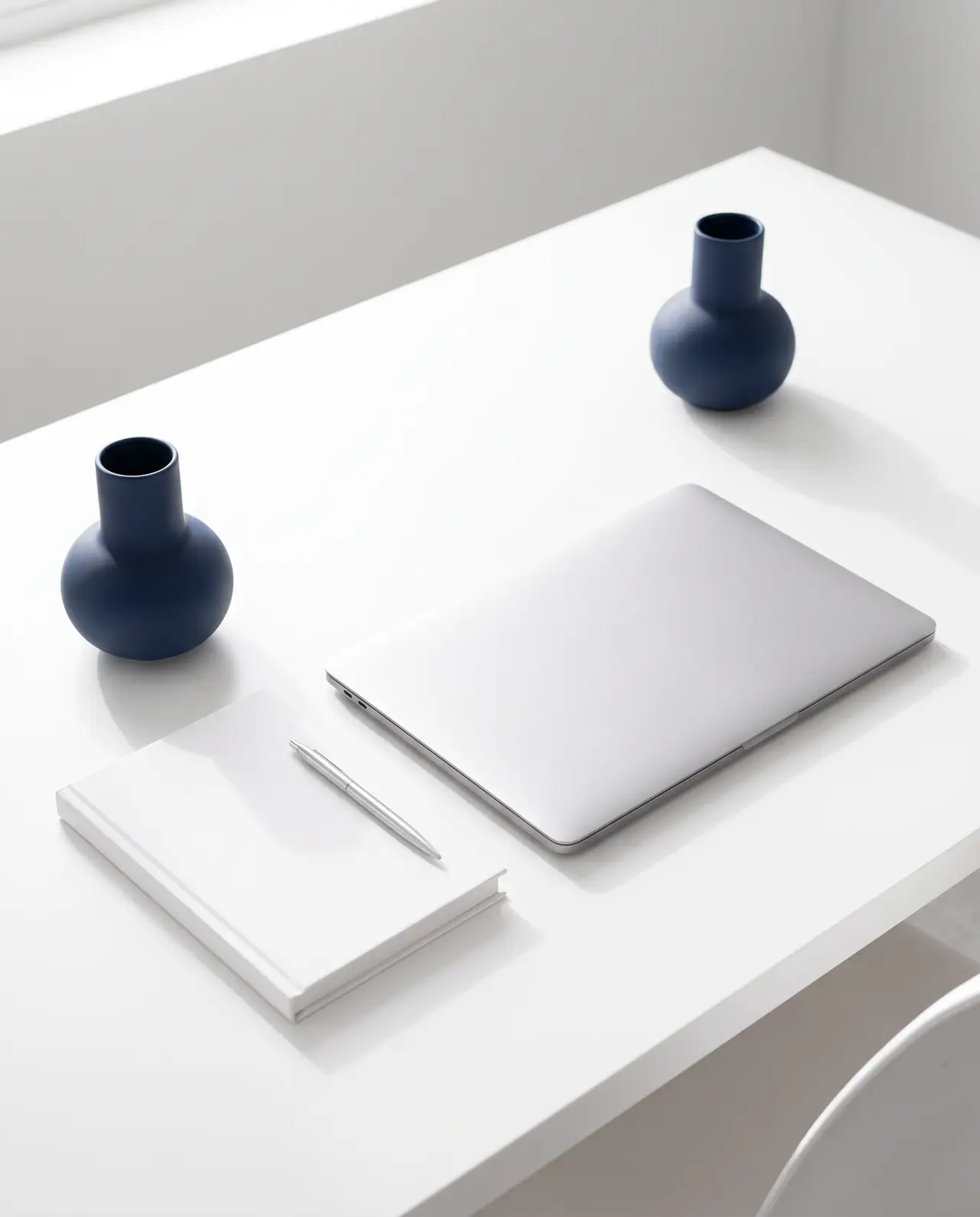 Mesa branca vista de cima com um notebook fechado, um caderno, uma caneta e dois vasos azul-marinho dispostos com espaço entre eles